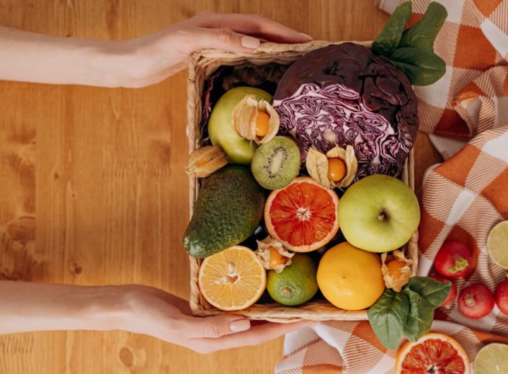 Vibrant basket of fresh fruits and vegetables with hands holding on a wooden table.